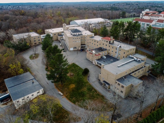 Aerial view of a large brick building complex with Caesar's Roofing, surrounded by trees, winding roads, grassy areas, and several parking lots; additional buildings and athletic fields are visible in the background.