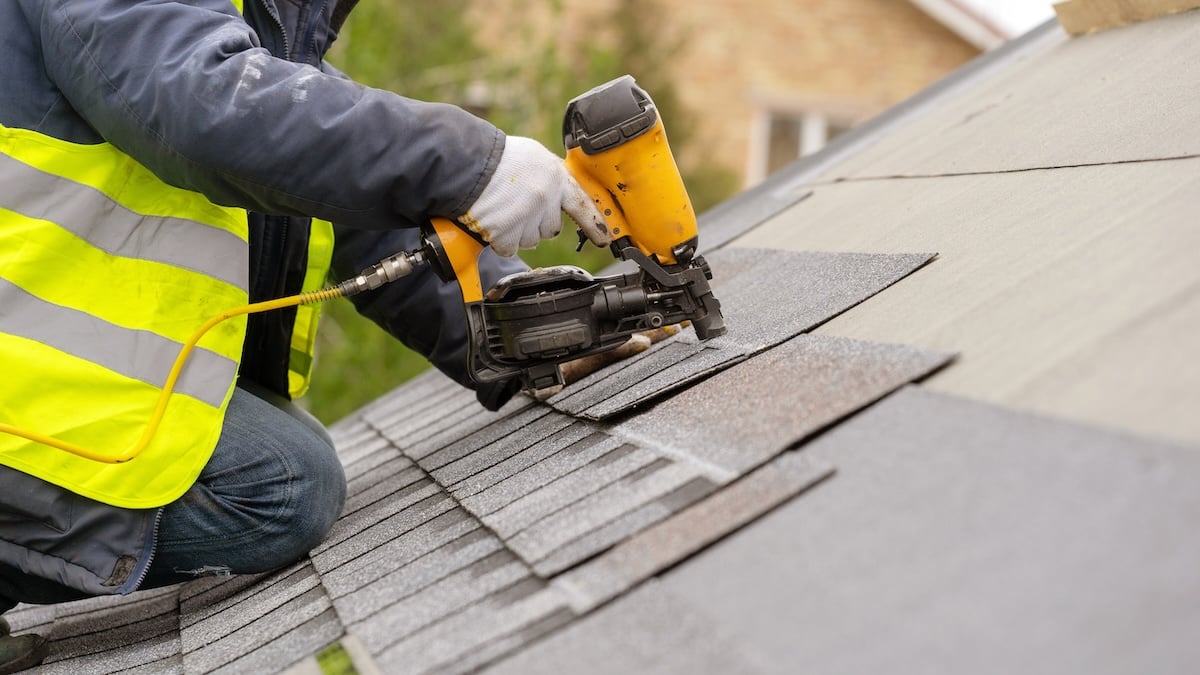 A worker wearing a high-visibility vest uses a nail gun to install asphalt shingles on a sloped roof. The worker’s hands are gloved and home exteriors are visible in the blurry background.