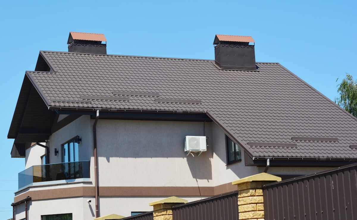 A modern house with a brown tiled roof, two large square chimneys, cream-colored walls, a glass balcony, and an outdoor air conditioning unit, set against a clear blue sky.
