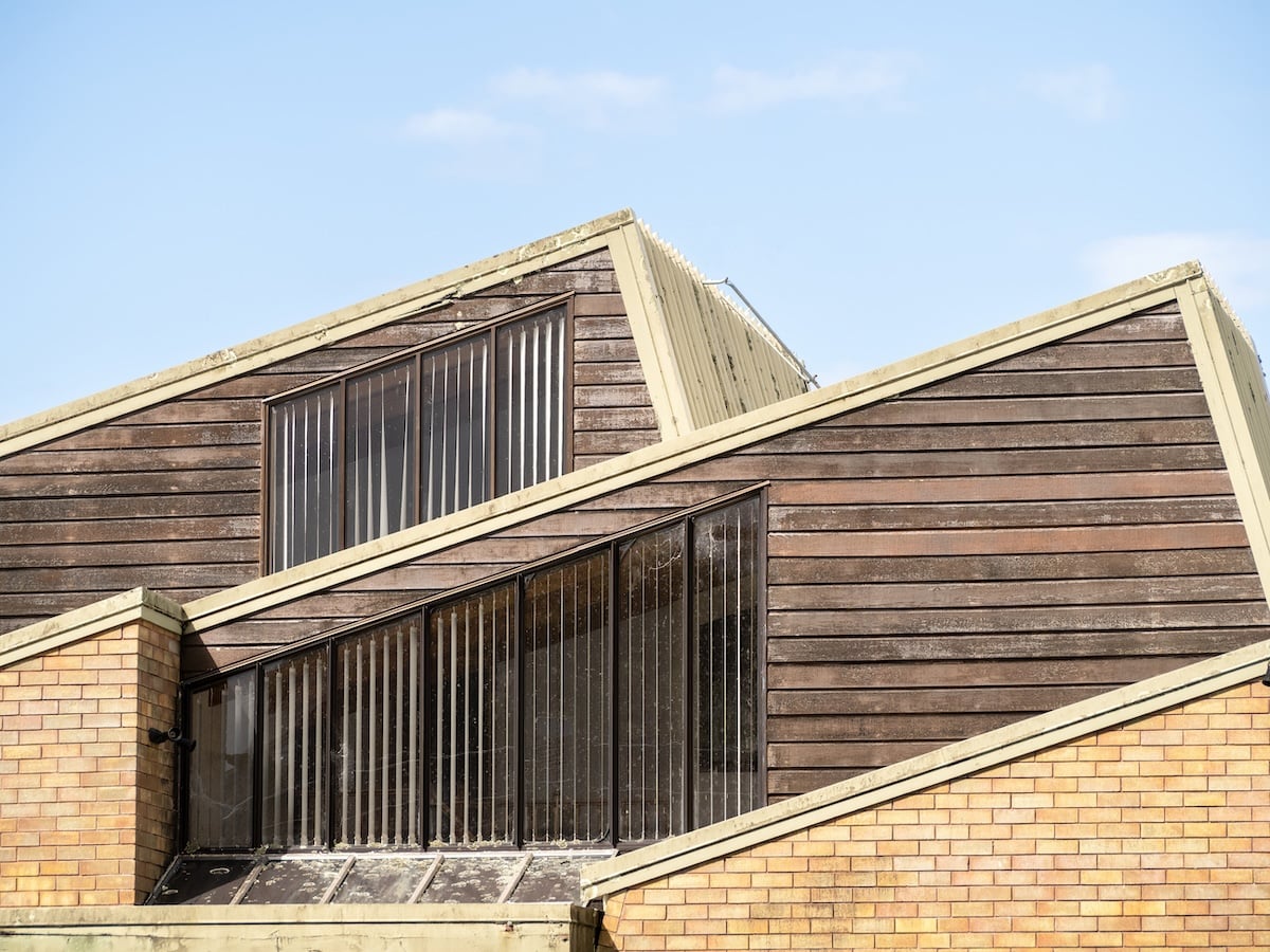 A building with modern, angular architecture featuring overlapping sloped roofs, wooden siding, large vertical windows, and a section of yellow brick under a clear blue sky.