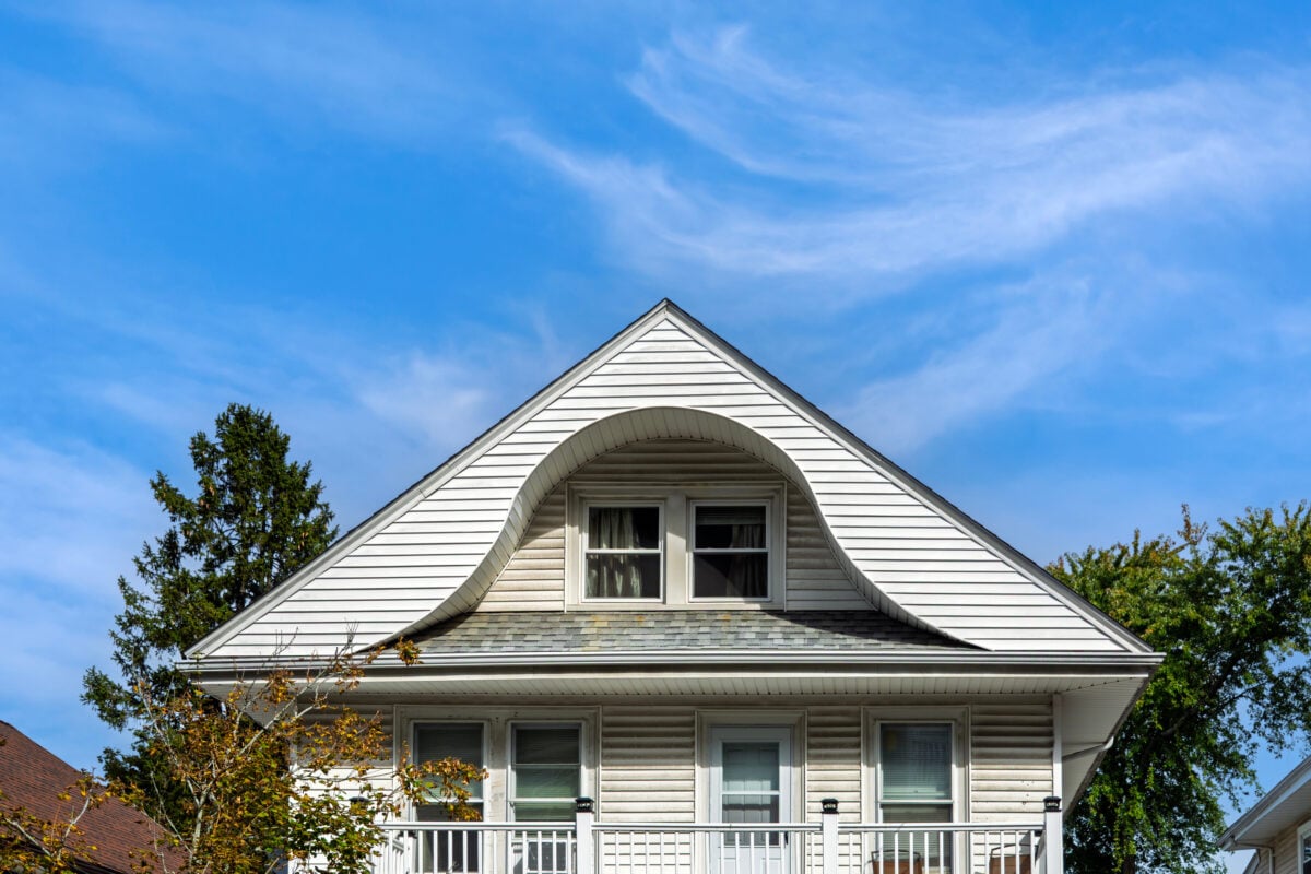 dormer roof White two-story house with a unique curved gable above the upper windows. The house has a balcony, siding, and is set against a blue sky with wispy clouds and some trees in the background.