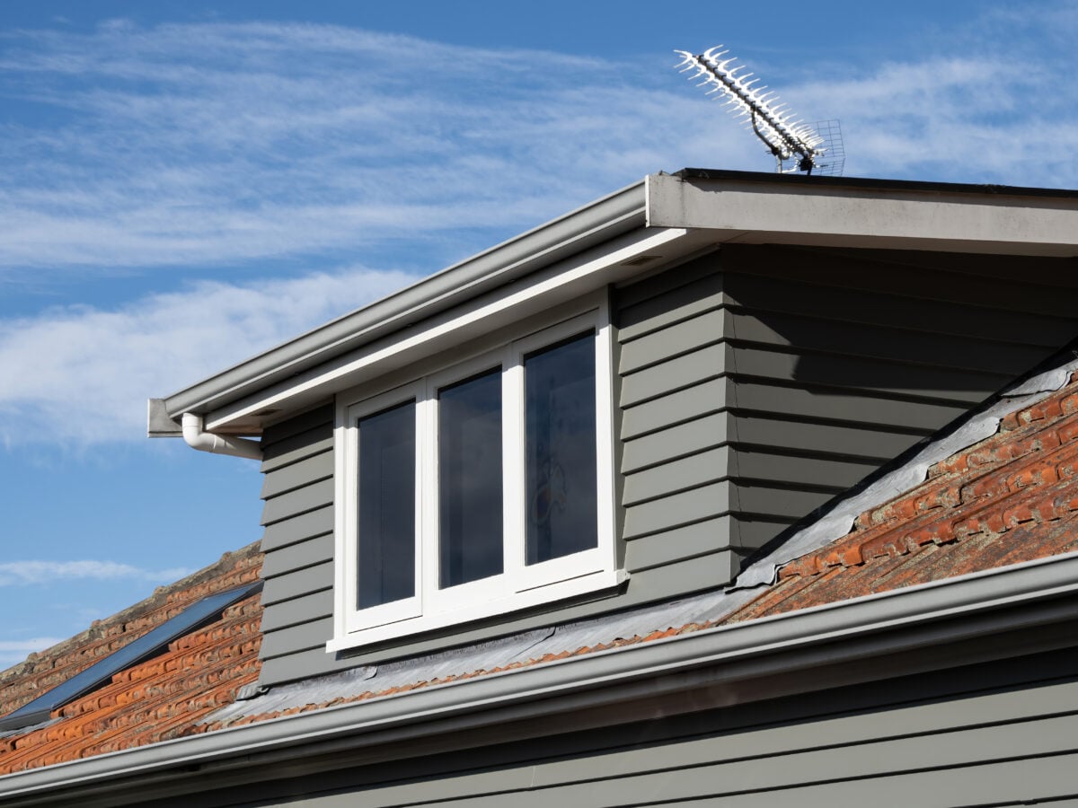 dormer roof A close-up of a house roof with gray horizontal siding, a white-framed window, and a TV antenna on top. The sky is blue with scattered clouds in the background.