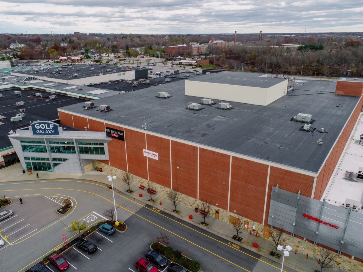 reliable roofers Aerial view of a large shopping mall with stores including Golf Galaxy and Sportsman's Warehouse, a parking lot with cars, and surrounding trees under a cloudy sky.