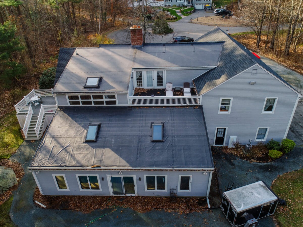 reliable roofers Aerial view of a large house with a flat roof section, skylights, a balcony with outdoor furniture, and a surrounding leaf-covered driveway. Trees with sparse leaves and neighboring houses are visible in the background.