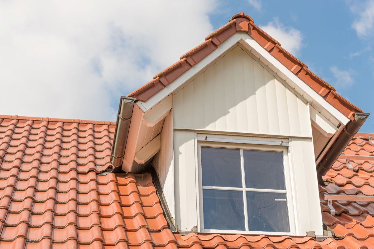 dormer roof A dormer window with white siding and a cross-pane design is set into a red, tiled sloped roof under a partly cloudy blue sky.