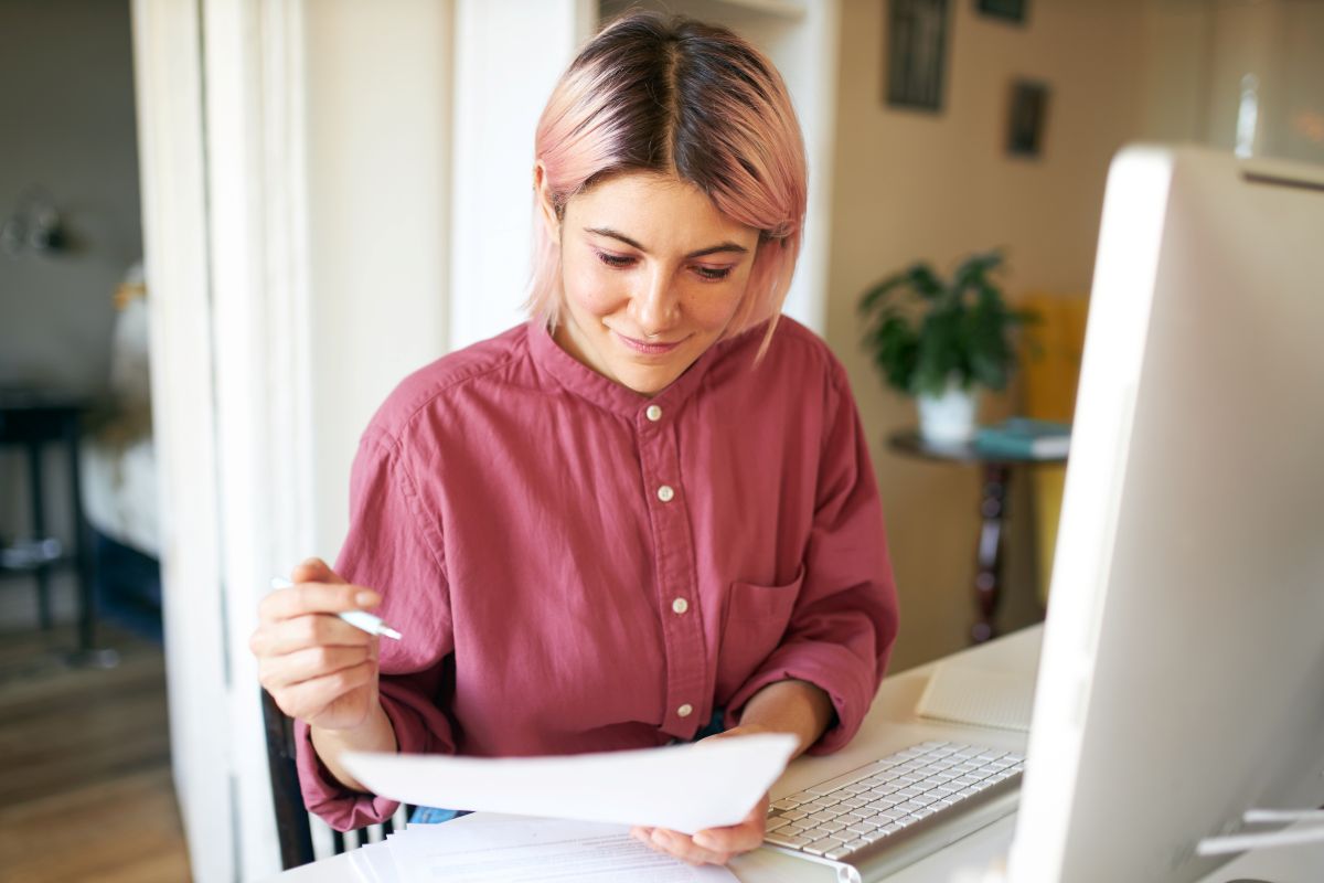 reliable roofers A person with short, pink hair and a maroon shirt sits at a desk, looking at a document while holding a pen. In front of them is an open laptop and a desktop monitor, in a brightly lit room.