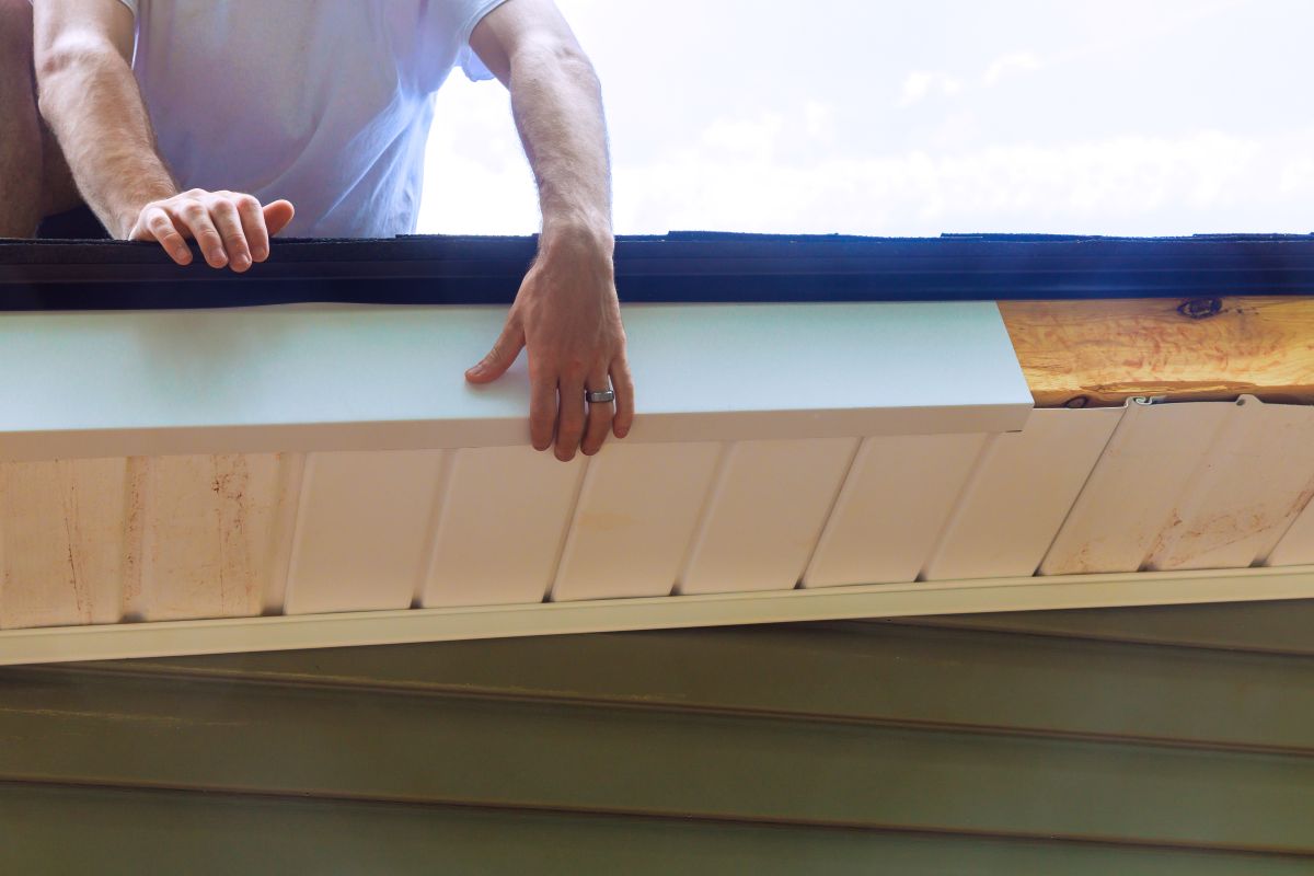 fascia board replacement A person installs a white soffit panel under the edge of a roof, with their hands securing the panel in place. The house has green siding and exposed wood near the roofline.