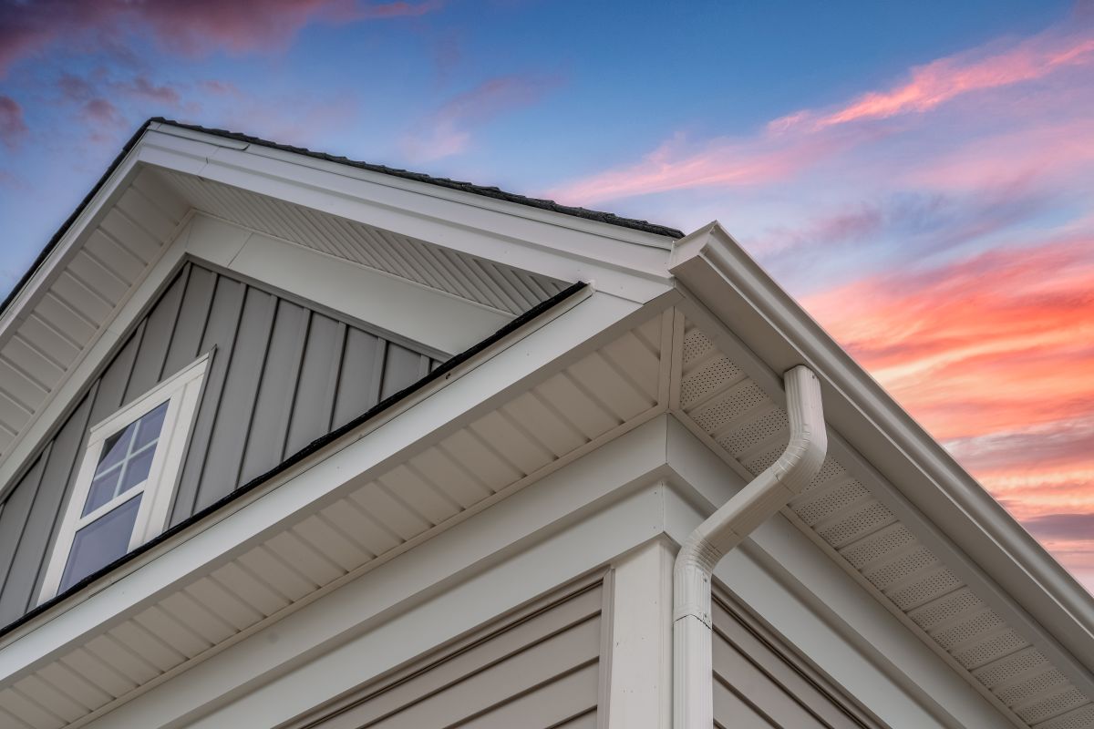 fascia board replacement Close-up view of a modern house’s roofline, gutter, and siding, with a window visible on the upper left. The sky in the background is colorful with shades of pink, orange, and blue at sunset.