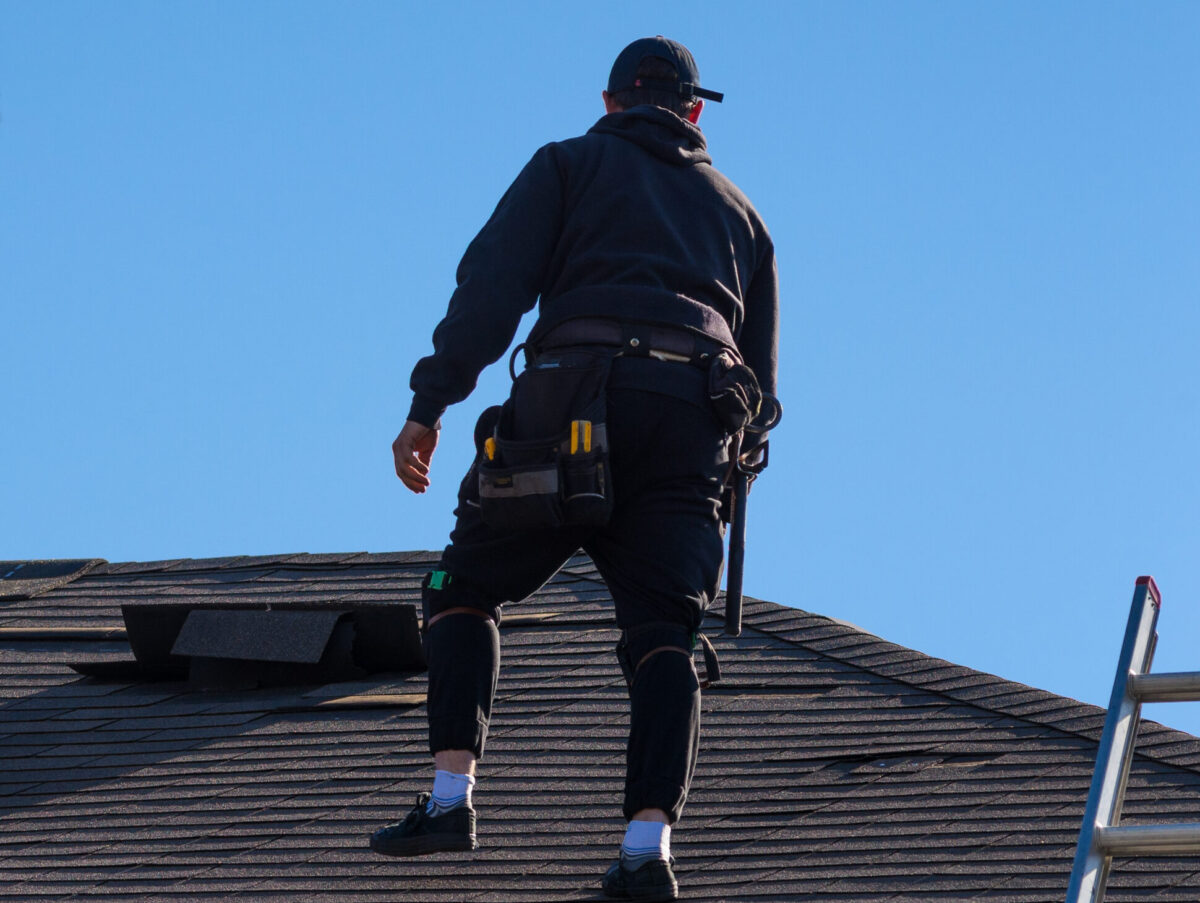 how to determine age of roof for insurance A roofer wearing a dark hoodie, cap, and tool belt stands on a shingled roof against a bright blue sky, with a metal ladder visible to the right.