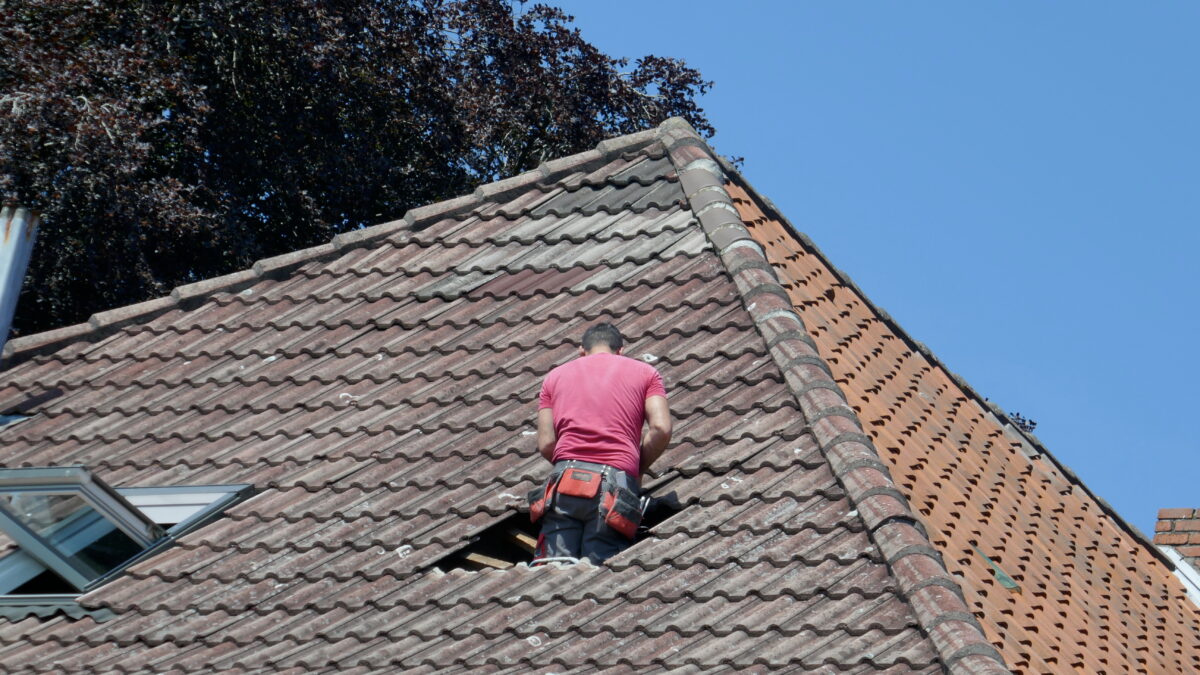 will insurance cover a 20 year-old roof A person wearing a red shirt and tool belt is standing on a tiled roof, facing an open window or hatch, with a clear blue sky and some tree branches visible in the background.