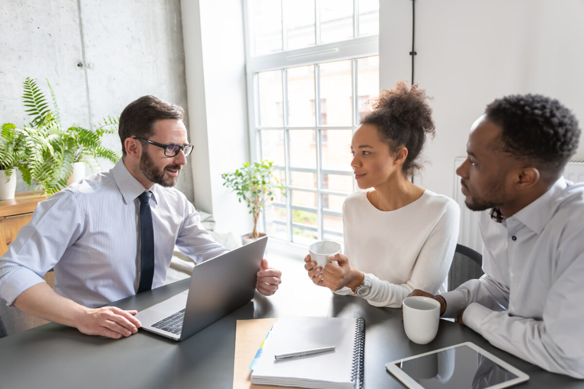Family couple consultations with a lawyer or insurance agent. Law and insurance. Three colleagues sit at a table with a laptop, notebooks, and coffee mugs, engaged in a discussion in a bright office with large windows and green plants.