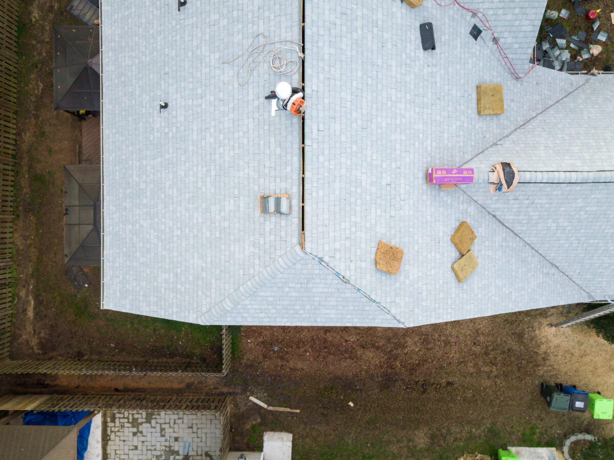 ridge vents Aerial view of a light gray shingled roof under construction, with tools, pink shingles, hoses, and some workers present. Brown patches and construction materials are visible on the ground below.