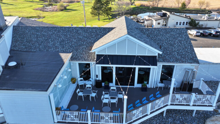 Residential Roofing Aerial view of a building with a deck featuring chairs and tables, string lights, and a surrounding outdoor area with green grass and parked cars.