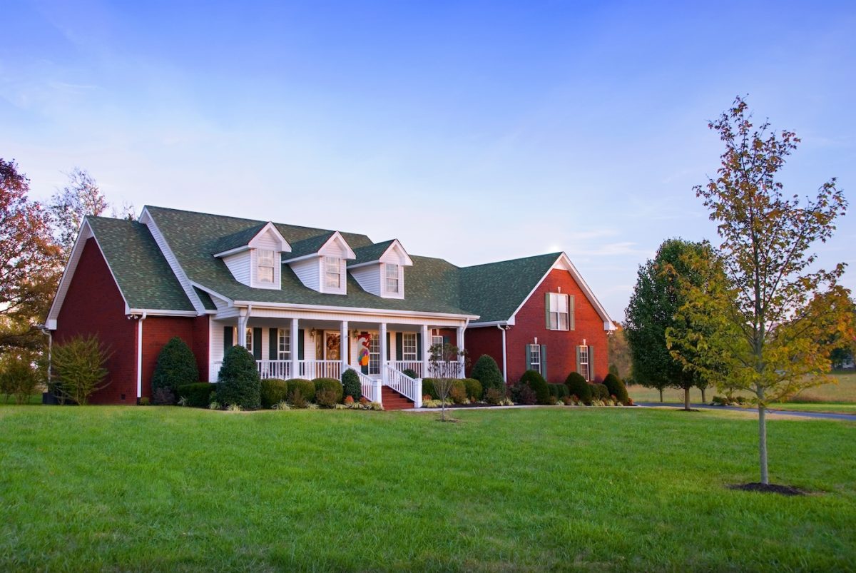 A large red brick house with white trim, a covered front porch, and green roof. It sits on a spacious, well-kept lawn with trees and shrubs, under a blue sky.