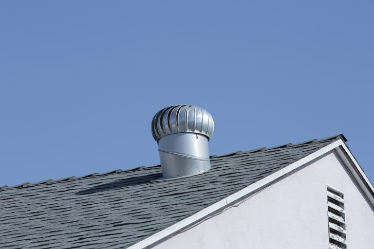 A metal turbine vent is installed on a gray shingled roof, set against a clear blue sky.